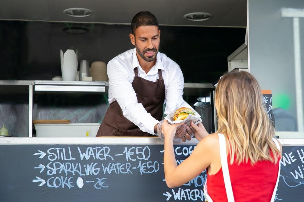 A person inside a food truck serves a hamburger to a customer while smiling