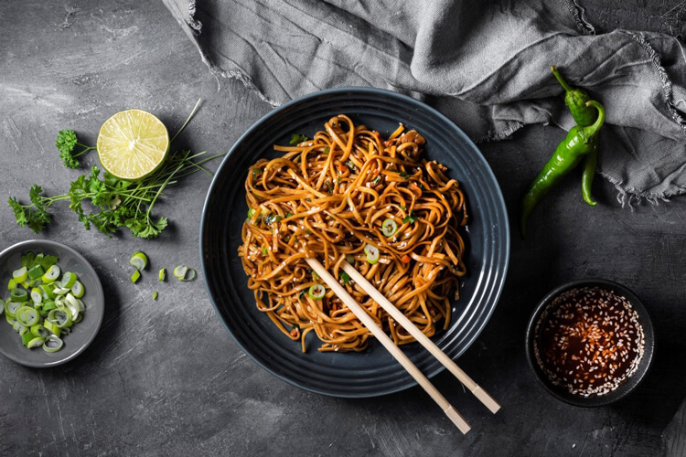 A black bowl of noodles with chopsticks and surrounded by ingredients