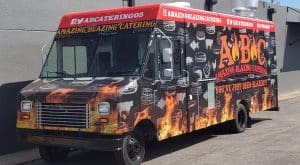 Amazing Blazing Catering food truck with a black wrap featuring realistic flame graphics along the lower body, white food and animal silhouettes, bold ABC logo, "You've Just Been Blazed" tagline, red header stripe with social media handles, and phone number
