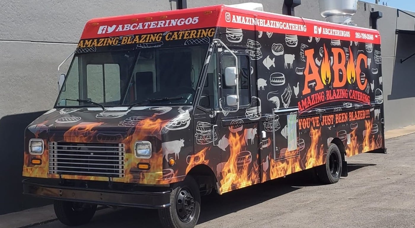 Amazing Blazing Catering food truck with a black wrap featuring realistic flame graphics along the lower body, white food and animal silhouettes, bold ABC logo, "You've Just Been Blazed" tagline, red header stripe with social media handles, and phone number