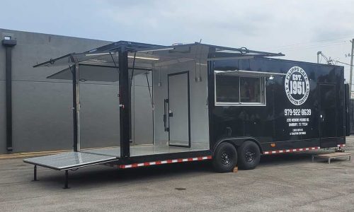 Black food trailer for 773 Vmo BBQ with a sliding service window, side porch extension, security lights, and branded door decal featuring a pitmaster graphic and QR code, parked at the JRS office