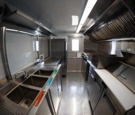 Wide-angle interior view of a custom food truck showing a three-compartment sink with Wash, Rinse, and Sanitize labels on the left, a full-height reach-in refrigerator at center, and a commercial cooking line with exhaust hood and white cutting board surface on the right, with diamond-plate flooring throughout