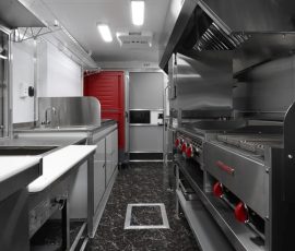 High-end food trailer interior featuring stainless steel prep counters with a white cutting board surface, commercial range with red control knobs, flat-top griddle, exhaust hood, hand sink, microwave, red reach-in refrigerator, exit sign, and dark marble-style flooring
