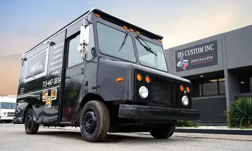 Matte black Kale's Kitchen food truck with branded wrap featuring menu item photos and contact number, parked outside JRS Custom Inc in Texas at dusk