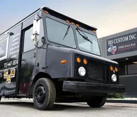 Matte black Kale's Kitchen food truck with branded wrap featuring menu item photos and contact number, parked outside JRS Custom Inc in Texas at dusk