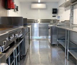 Food truck interior showing stainless steel prep tables and cooking equipment on both sides, a mini-split AC unit, fire suppression system, hand sink, paper towel dispenser, and polished diamond-plate flooring