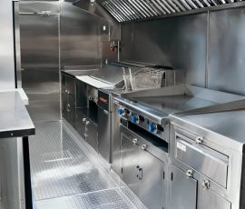 Interior of a custom food truck kitchen with full stainless steel construction, including a commercial flat-top griddle, deep fryer with basket, exhaust hood, under-counter storage cabinets, and diamond-plate flooring