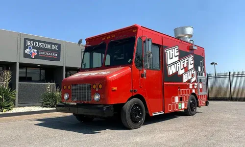 Bright red The Waffle Bus food truck with bold black and white branding and a geometric pattern along the lower body, parked outside JRS Custom Inc in Texas