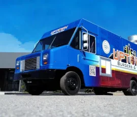 Uptown Burgers and Fish food truck with a vivid blue and red gradient wrap, city skyline graphic, QR code, and phone number, shot from a low angle against a clear blue sky