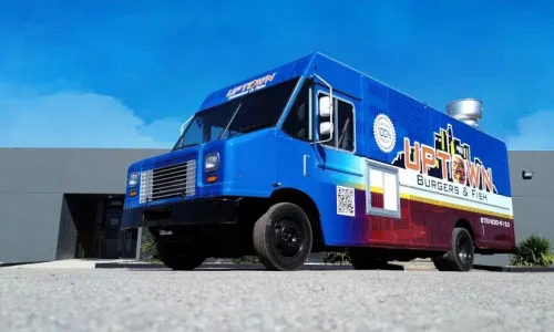Uptown Burgers and Fish food truck with a vivid blue and red gradient wrap, city skyline graphic, QR code, and phone number, shot from a low angle against a clear blue sky