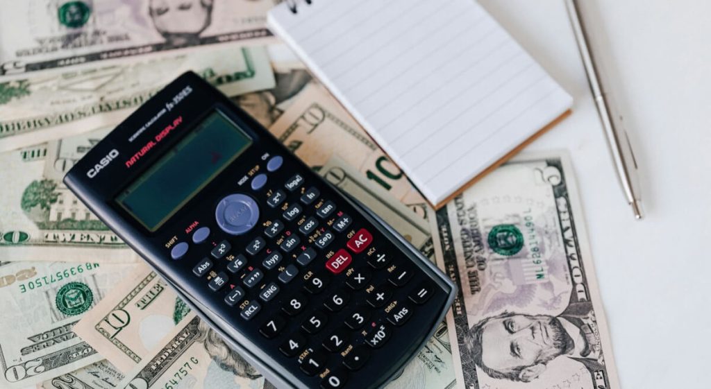 Calculator resting on top of cash bills next to a notepad and pen