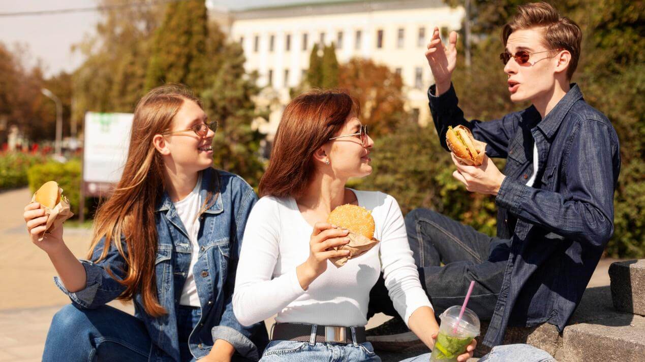 Three friends sitting on the floor with burgers in their hands, chatting and smiling