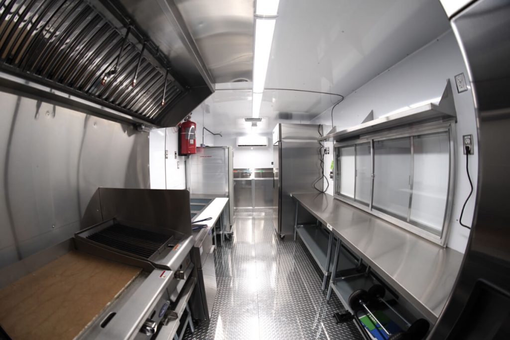 Interior of a custom food truck build showing stainless steel kitchen layout with charbroiler, commercial refrigerator, prep counters, ventilation hood, and service window