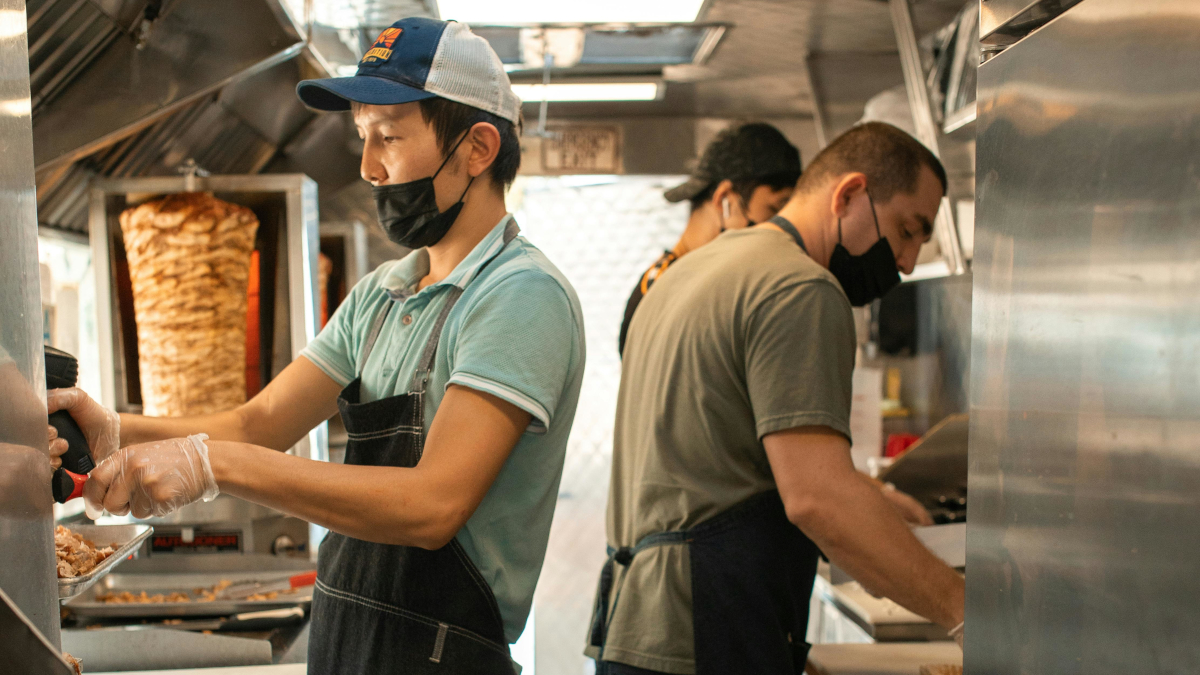 Three food truck cooks preparing orders in a stainless steel kitchen with a trompo spit in the background