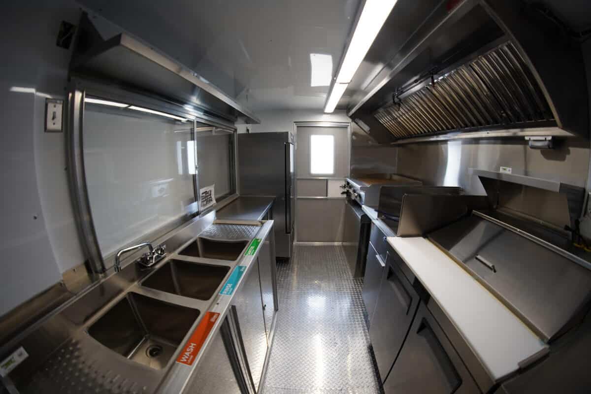 Wide-angle interior view of a custom food truck showing a three-compartment sink with Wash, Rinse, and Sanitize labels on the left, a full-height reach-in refrigerator at center, and a commercial cooking line with exhaust hood and white cutting board surface on the right, with diamond-plate flooring throughout