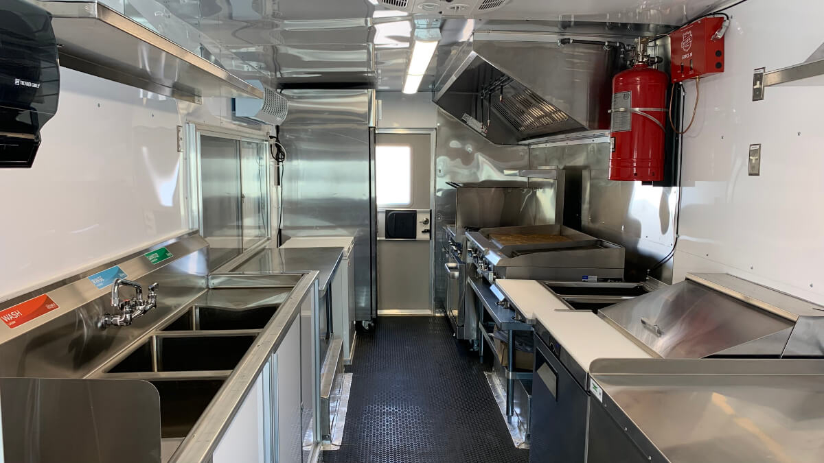Stainless steel food truck kitchen interior with a commercial cooking line, exhaust hood, three-compartment sink, and LED lighting