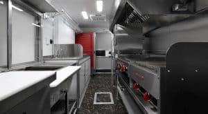 High-end food trailer interior featuring stainless steel prep counters with a white cutting board surface, commercial range with red control knobs, flat-top griddle, exhaust hood, hand sink, microwave, red reach-in refrigerator, exit sign, and dark marble-style flooring