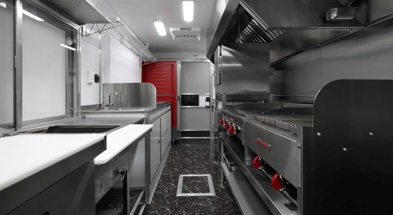 High-end food trailer interior featuring stainless steel prep counters with a white cutting board surface, commercial range with red control knobs, flat-top griddle, exhaust hood, hand sink, microwave, red reach-in refrigerator, exit sign, and dark marble-style flooring