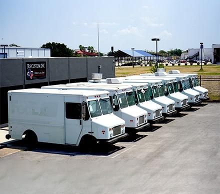 A row of eight white step van food trucks lined up in the lot outside JRS Custom Inc, the Jerusalem Custom Food Trucks & Trailers fabrication facility in Texas