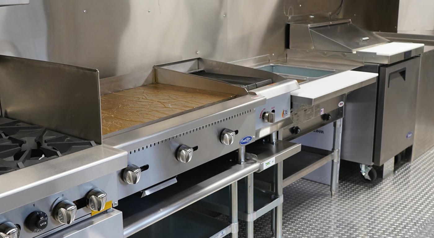 Commercial food truck cooking line showing a gas range, CookRite flat-top griddle, deep fryer, and under-counter refrigerator unit lined up on diamond-plate flooring