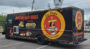 La Lupe Mexican Food truck with a black wrap featuring a large circular logo with a traditional Mexican woman mascot, food photos of tacos, quesadillas, burritos, and tortas, gold food icon border pattern, and to-go order phone number
