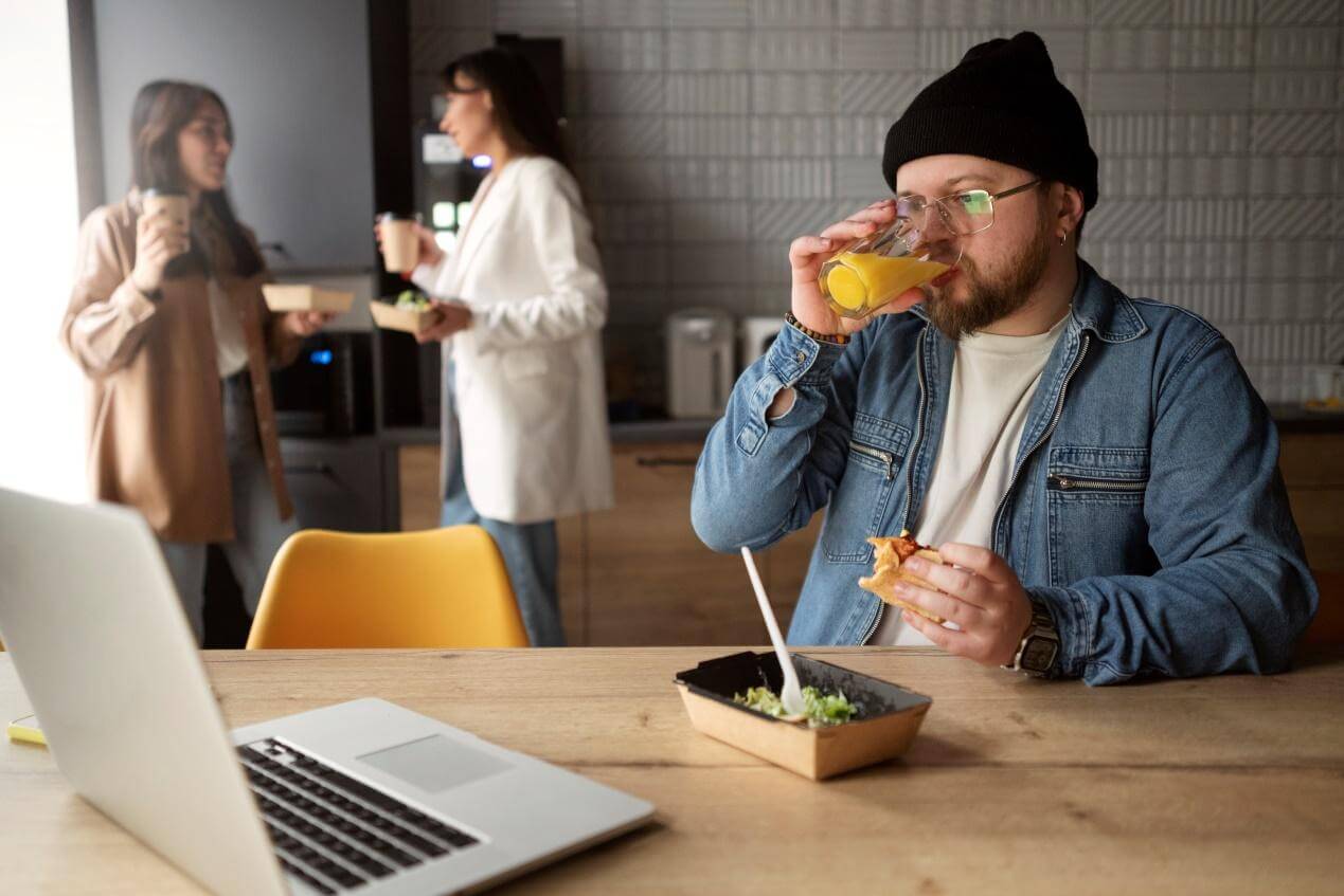 A young man sitting on a chair, eating food and drinking juice while looking at the laptop screen