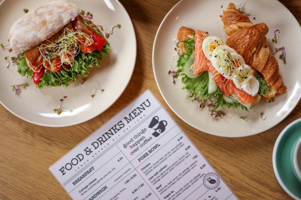 Overhead view of two plated sandwiches beside a printed food and drinks menu on a wooden table