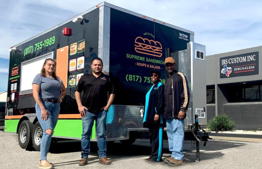 Four people posing in front of the completed Supreme Sandwiches food trailer showing the rear wrap with branding, logo, and contact information, outside JRS Custom Inc in Texas
