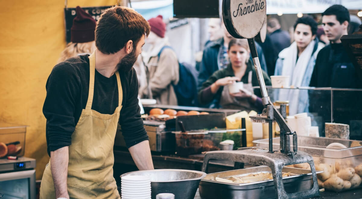 Food truck cook preparing orders at a busy outdoor market service counter with customers in line