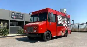 Bright red The Waffle Bus food truck with bold black and white branding and a geometric pattern along the lower body, parked outside JRS Custom Inc in Texas