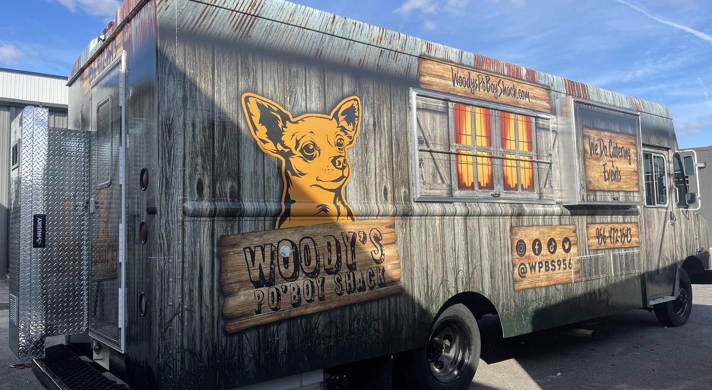 Woody's Po'Boy Shack food truck with a rustic weathered wood plank wrap featuring a Chihuahua mascot illustration, barn window graphic, catering callout, social media handles, phone number, and diamond-plate exterior storage compartment